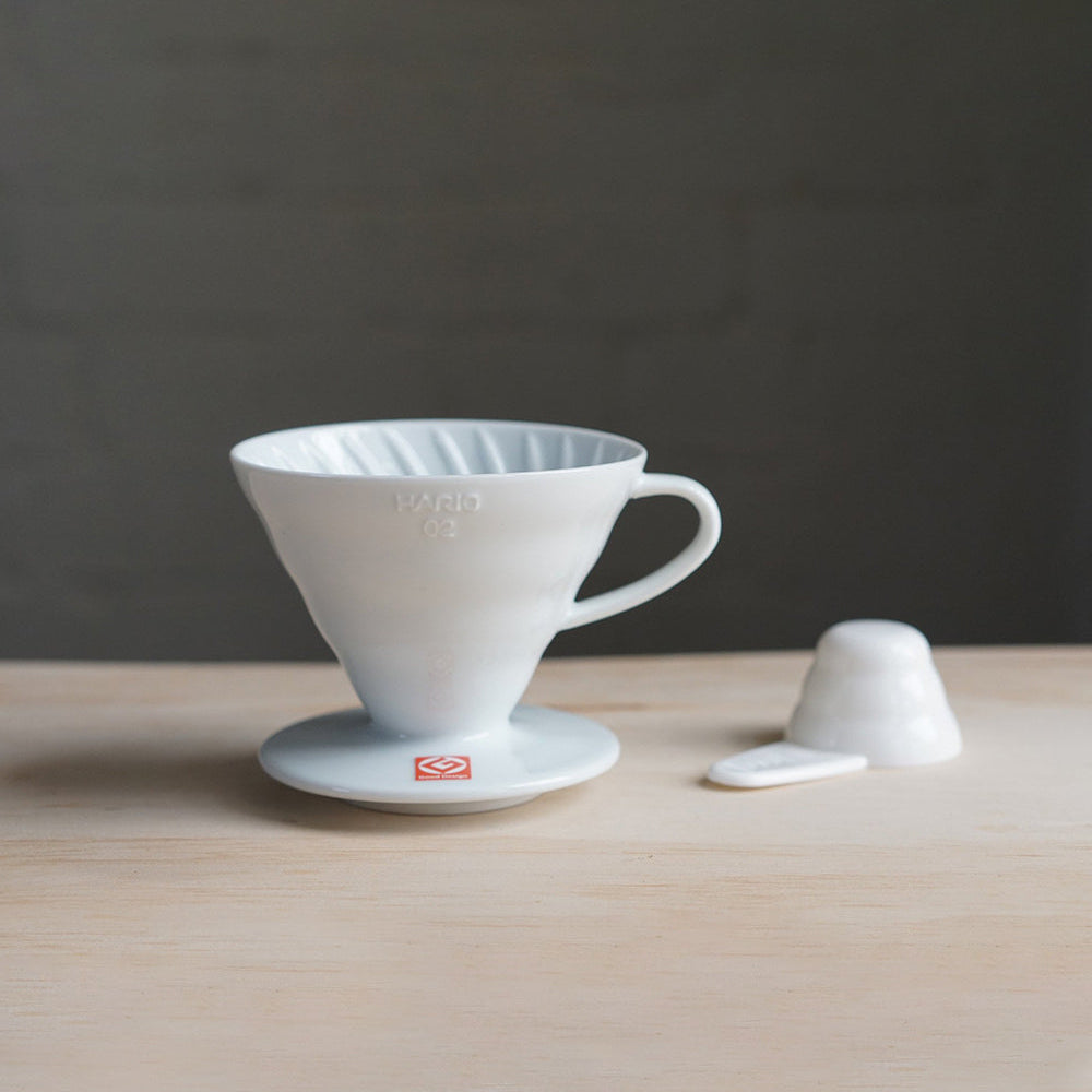 A white ceramic Hario V60 coffee dripper with a cup and a small white paper filter on a wooden surface.