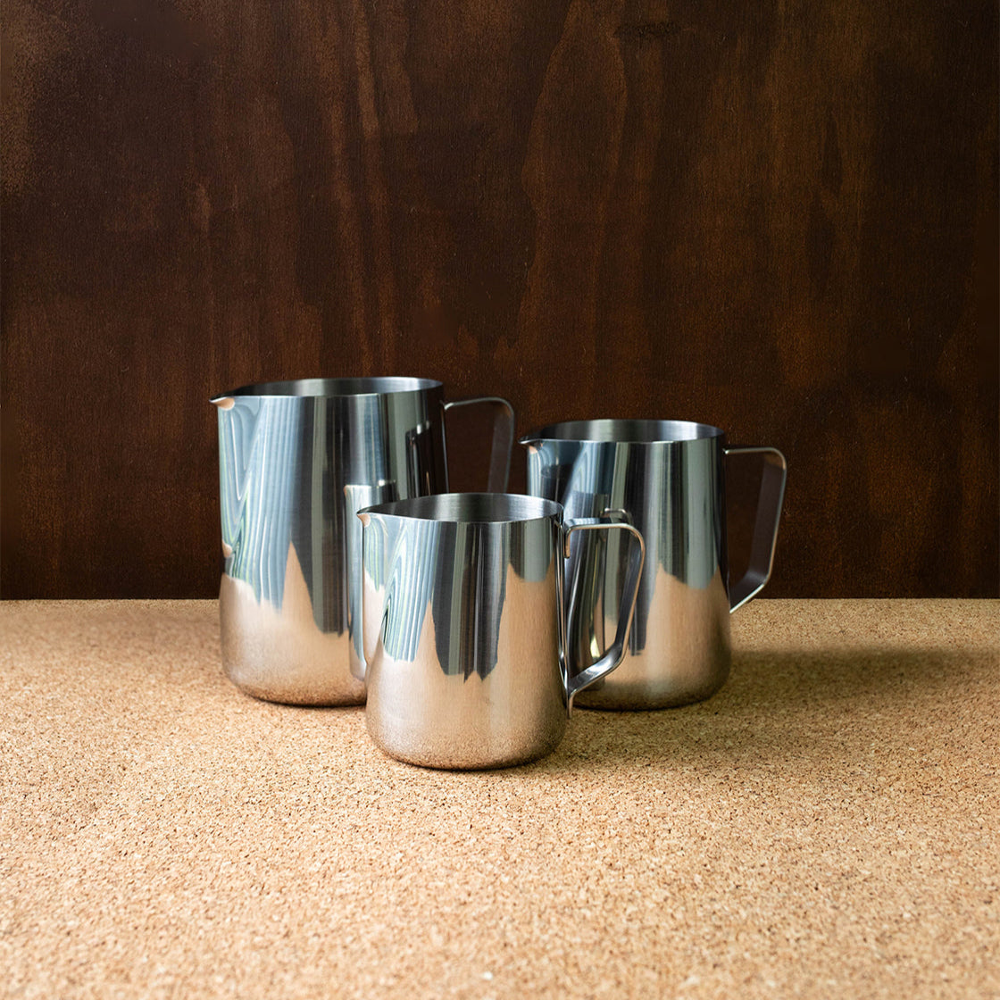 Three stainless steel milk jugs of different sizes arranged on a surface, likely intended for making and serving steamed milk in coffee beverages.